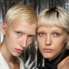 Zwei Models mit kurzen, blonden Haaren und blassen Gesichtern vor silbrigem Hintergrund.