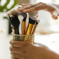 Hände halten einen goldenen Behälter mit verschiedenen Make-up-Pinseln.