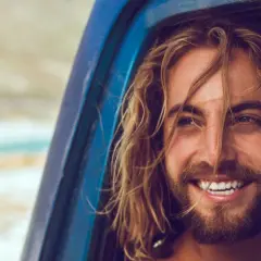Mann mit langen, welligen Haaren und Bart lächelt aus einem blauen Auto.