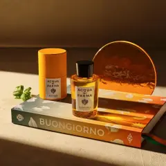 Acqua di Parma Flakon und Dose auf Buch mit "Buongiorno" Schriftzug, daneben ein grüner Zweig.
