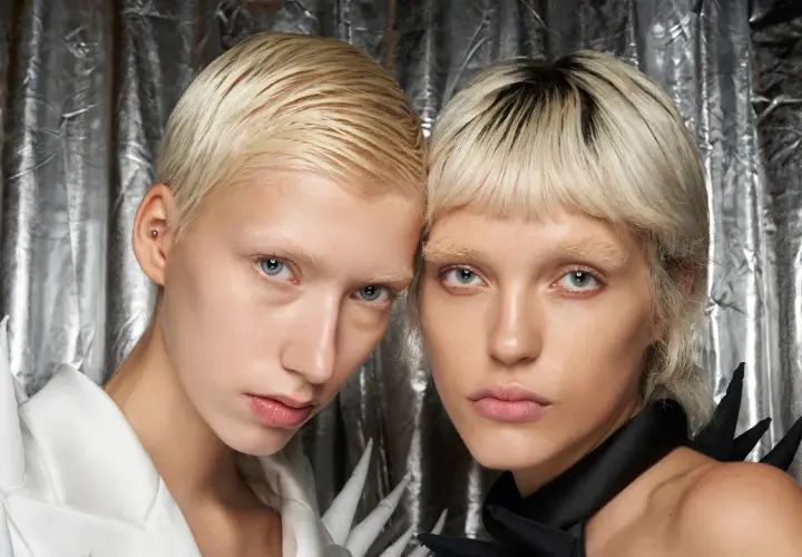 Zwei Models mit kurzen, blonden Haaren und blassen Gesichtern vor silbrigem Hintergrund.