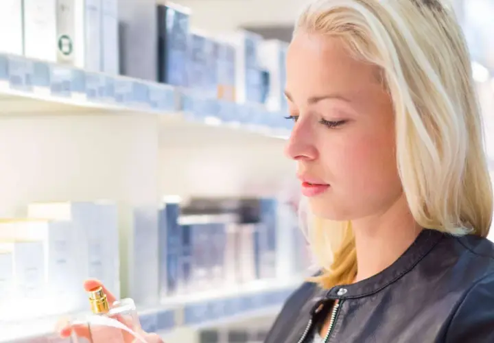 Blonde Frau mit Parfumflakon in der Hand vor einem Regal mit Kosmetikprodukten.