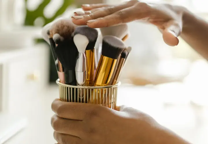 Hände halten einen goldenen Behälter mit verschiedenen Make-up-Pinseln.