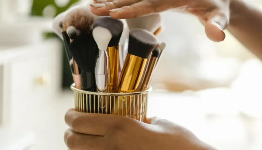 Hände halten einen goldenen Behälter mit verschiedenen Make-up-Pinseln.