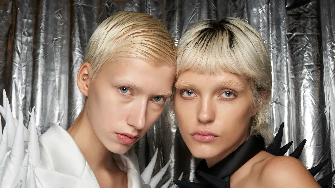 Zwei Models mit kurzen, blonden Haaren und blassen Gesichtern vor silbrigem Hintergrund.