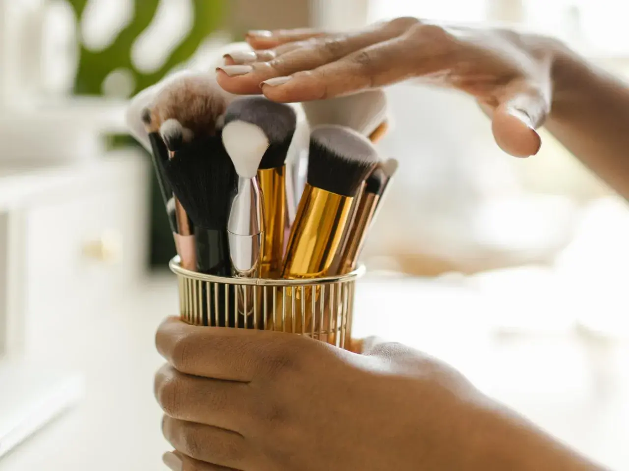 Hände halten einen goldenen Behälter mit verschiedenen Make-up-Pinseln.
